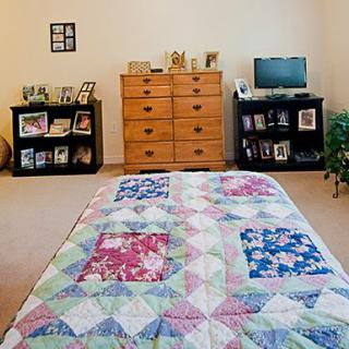 A cozy bedroom featuring a colorful quilt on the bed, a wooden dresser, and shelves with various decorations.