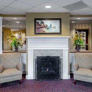 A cozy lobby interior featuring a fireplace, two chairs, decorative flowers, and framed artwork.