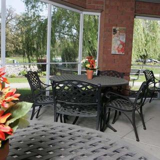 An outdoor patio with iron furniture and colorful flower arrangements, overlooking a scenic garden through large glass windows.