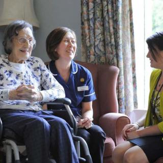 A caregiver and a nurse engage in conversation with an elderly woman in a wheelchair.