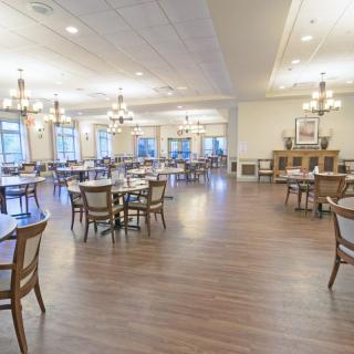 A spacious dining room with wooden flooring, featuring multiple tables and chairs, under elegant lighting.