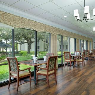 A well-lit dining area with large windows, featuring wooden floors and a view of the greenery outside.