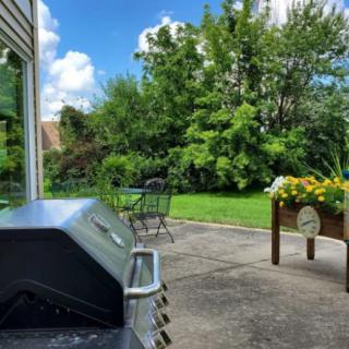 A sunny backyard patio with a grill and seating area surrounded by lush greenery.
