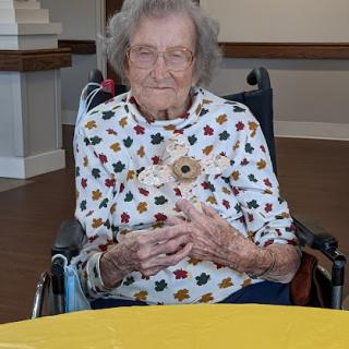 An elderly woman in a wheelchair, wearing a colorful polka dot shirt, smiles warmly while sitting at a table.