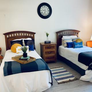 A cozy bedroom with twin beds, featuring colorful pillows and a central nightstand under a wall clock.