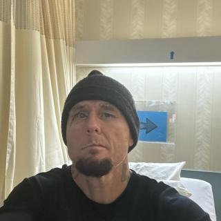 A man wearing a black beanie is taking a selfie in a room with curtains and a bed.
