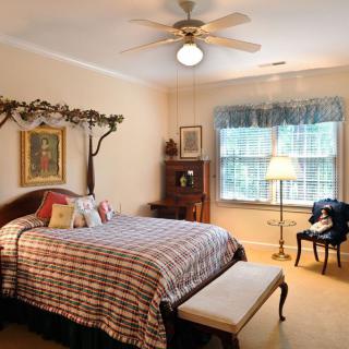 A traditional-style bedroom with a large bed, a wooden desk and chair, a lamp by the window, and decorative elements.