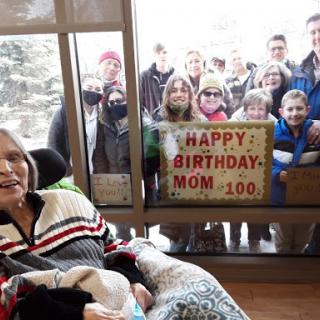 A joyful celebration of a mother's 100th birthday, with family gathered outside to send their love.