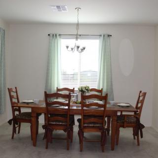 A well-lit dining room featuring a wooden table and chairs, with a chandelier overhead and curtains framing the window.
