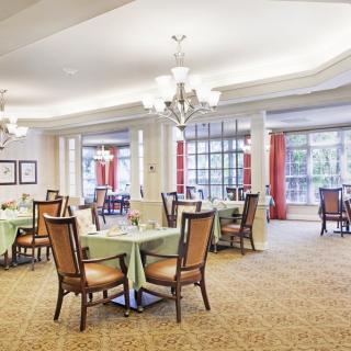 A spacious and elegantly decorated dining room with multiple tables, chairs, and chandeliers, illuminated by natural light from large windows.