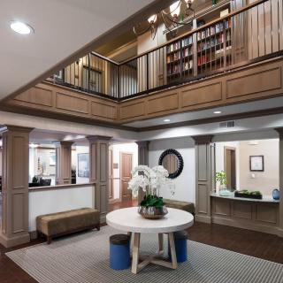 A modern lobby with a central round table, decorative plants, and an upstairs bookshelf area.