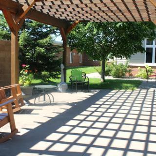 A serene outdoor space with a wooden pergola casting shadows on the ground, surrounded by lush greenery.