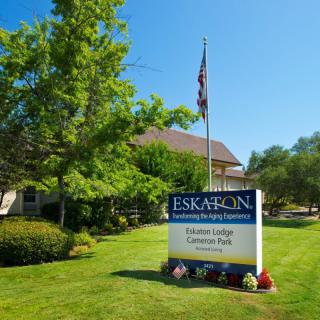 A sign for Eskaton Village Carmichael is displayed on a well-maintained lawn, accompanied by trees and an American flag on a sunny day.