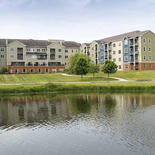 A serene view of modern apartments beside a calm lake, surrounded by lush greenery.