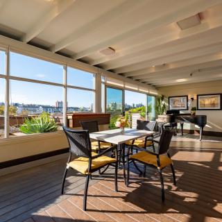 A modern dining area with large windows allowing natural sunlight, featuring a table set with chairs and a piano in the corner.