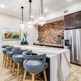 A modern kitchen featuring a large island with bar stools, pendant lighting, a stone backsplash, and stainless steel appliances.