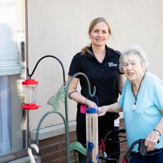 A caregiver assisting an elderly woman with bird feeders outside.