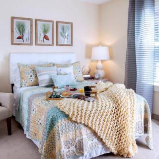 A cozy bedroom featuring a neatly made bed with patterned pillows and a chunky knit blanket, complemented by soft lighting from a table lamp and artwork above the headboard.