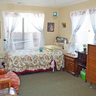 A cozy bedroom with a bed, dresser, and chair, featuring floral bedding and sheer curtains.