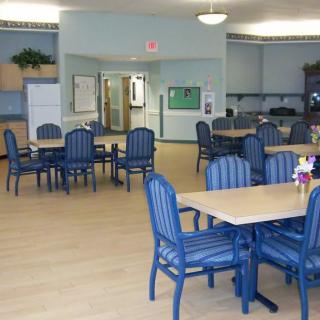 A well-lit dining area with blue chairs and wooden tables, featuring a kitchen in the background.