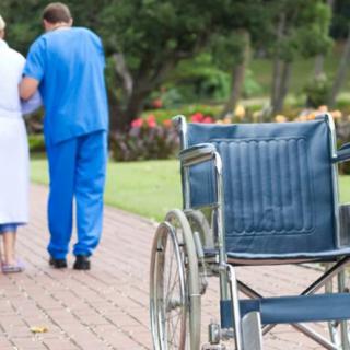 A caregiver assists an elderly person walking outdoors with an empty wheelchair nearby.