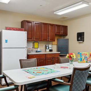 A cozy kitchen area with a table set for games and flowers, surrounded by cabinets and appliances.