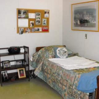 A cozy bedroom with a single bed, featuring a colorful quilt and decorative pillows. A bulletin board adorned with photos and notes is on the wall, and a small shelf holds various items.