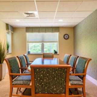 A well-lit conference room with a wooden table, surrounded by upholstered chairs, featuring a window and indoor plant.