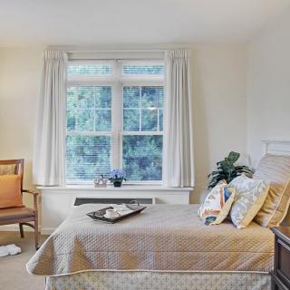 A cozy bedroom with a neatly made bed, a wooden chair beside a large window letting in natural light.