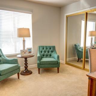 A cozy living room with two teal armchairs, a wooden side table, and a large mirror reflecting light from the window.