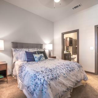 A cozy bedroom with a neatly made bed, two nightstands with lamps, and an en-suite bathroom visible through an open door.
