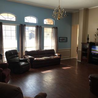 A spacious living room with large windows, leather sofas, a chandelier, and wooden flooring.