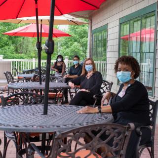 A group of individuals wearing masks are seated at an outdoor patio, practicing social distancing under red umbrellas.