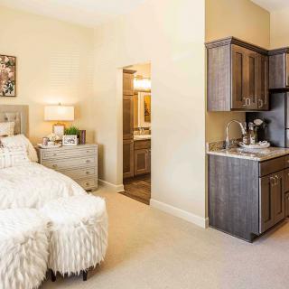 A cozy bedroom with a kitchenette, featuring a plush bed, side table, lamp, and decorative elements.