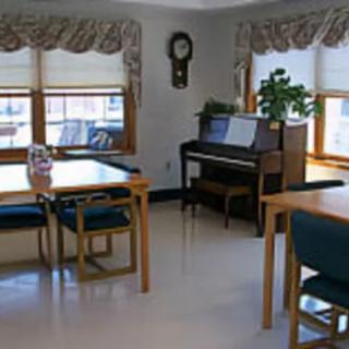 A cozy living room featuring a piano, tables, and chairs near large windows.