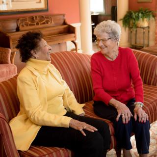Two elderly women sitting on a couch and engaging in a lively conversation.