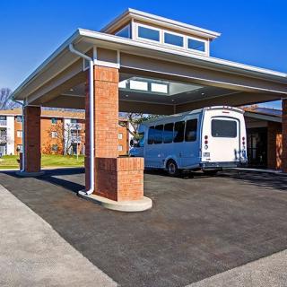A white bus parked under a canopy in front of a brick building on a sunny day.