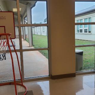 A view from inside a school showcasing a basketball hoop and the outdoor courtyard through large windows.