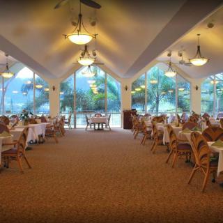 A spacious restaurant interior with neatly arranged tables and chairs, illuminated by elegant lighting fixtures and large windows offering a scenic view.