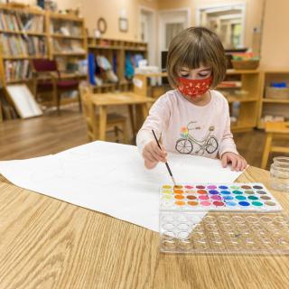 A young child wearing a mask is engaged in painting with watercolors at a table indoors.