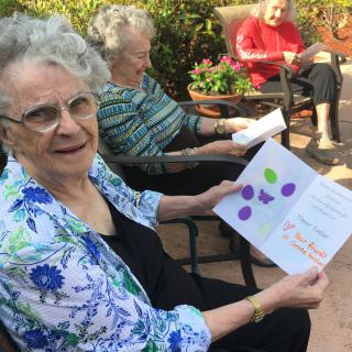 An elderly woman seated outdoors in a garden, holding and showing a card while others sit nearby.