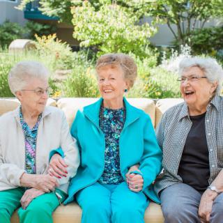 Three elderly women enjoying each other's company while sitting on a bench outdoors.