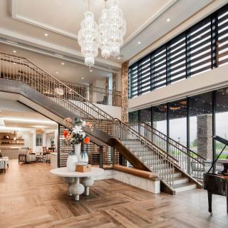 A luxurious lobby featuring a grand staircase, elegant chandelier, and a grand piano, with large windows providing ample natural light.