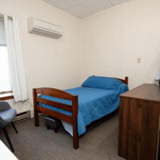 A small bedroom with a single bed covered in blue bedding, a wooden dresser, a chair by the window, and an air conditioning unit on the wall.