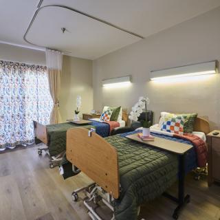A hospital room with two patient beds equipped with bedside tables and overhead lighting.