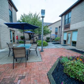 A cozy courtyard featuring outdoor seating with umbrellas, surrounded by brick pathways and lush greenery.