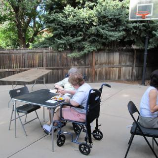 A person in a wheelchair sits at a table outdoors, next to another individual with a basketball hoop in the background.