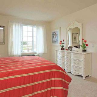 A cozy bedroom with a red bedspread, a dresser with a mirror, and natural light coming through a window.