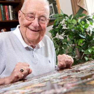 An elderly man smiles while working on a jigsaw puzzle at home.