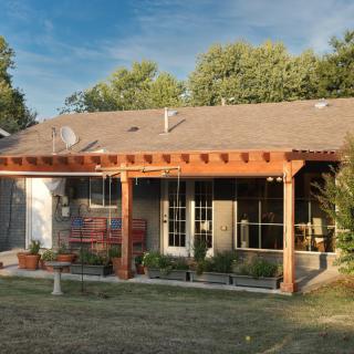 A backyard view of a house with a wooden pergola and patio area, surrounded by greenery and outdoor furniture.